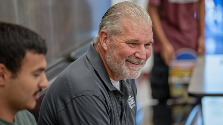 Two people sitting and smiling, with one person in focus. They're indoors near a table.