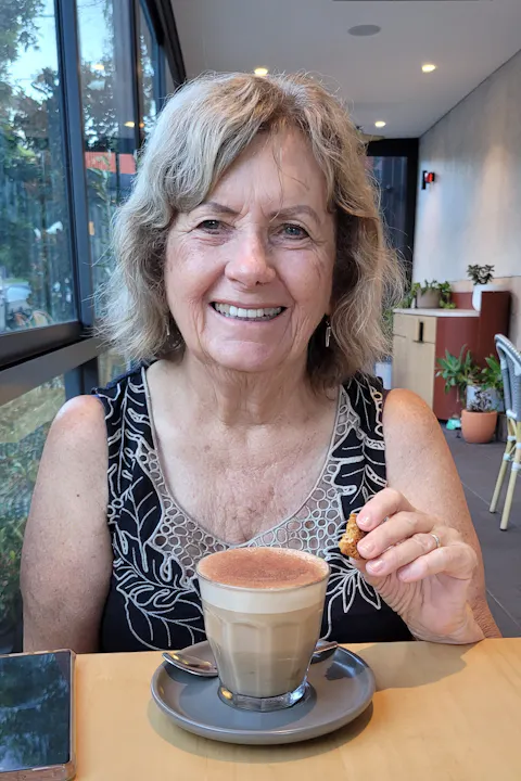 A smiling woman holds a cookie over a latte in a cozy café setting, with plants in the background.