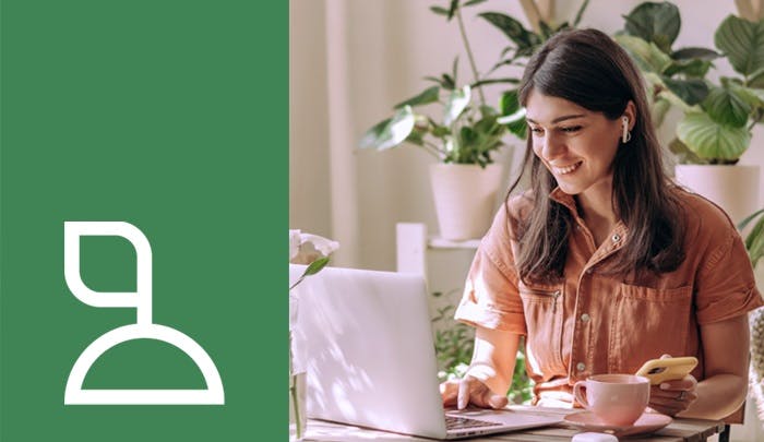 A smiling woman using a laptop with earphones, surrounded by plants, holding a cup and a smartphone.