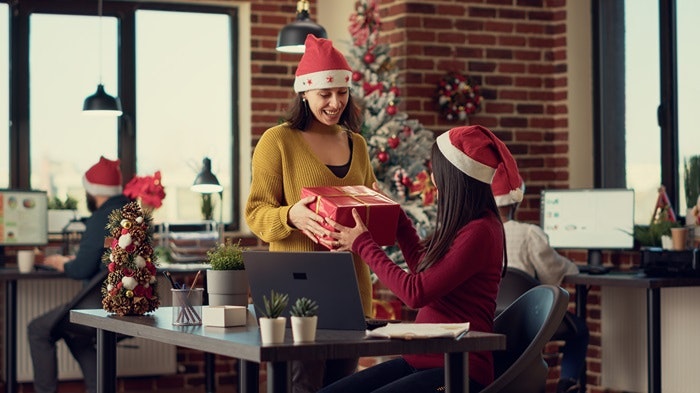 Two people wearing Santa hats exchange a gift in a festive office setting.