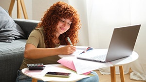 A person with curly hair is studying at a laptop, writing notes on papers, with a calculator and phone nearby.
