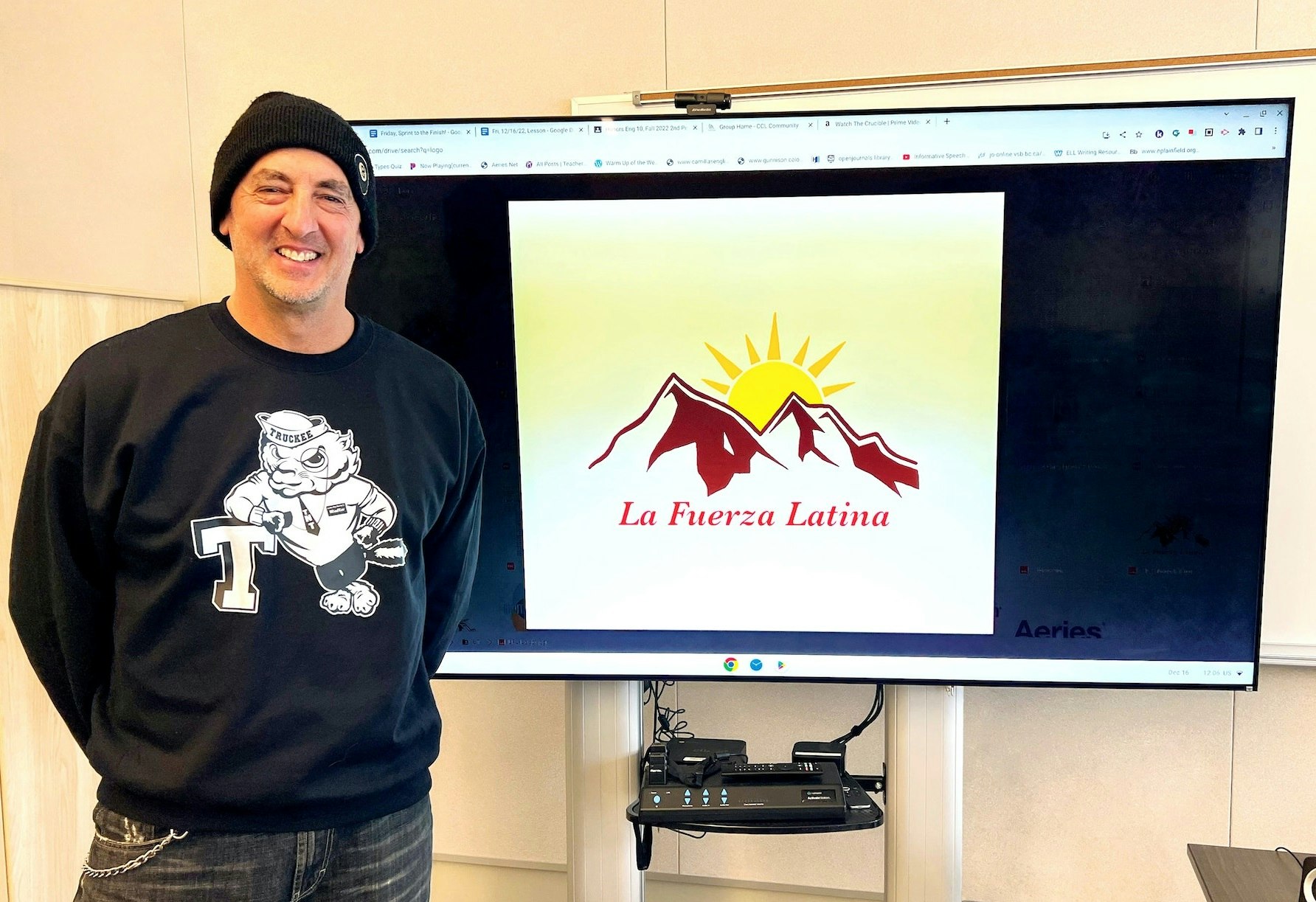 Craig Rowe wears a black stocking cap and a black sweatshirt and blue jeans. He is smiling and stands in front of a digital display for La Fuerza Latina.