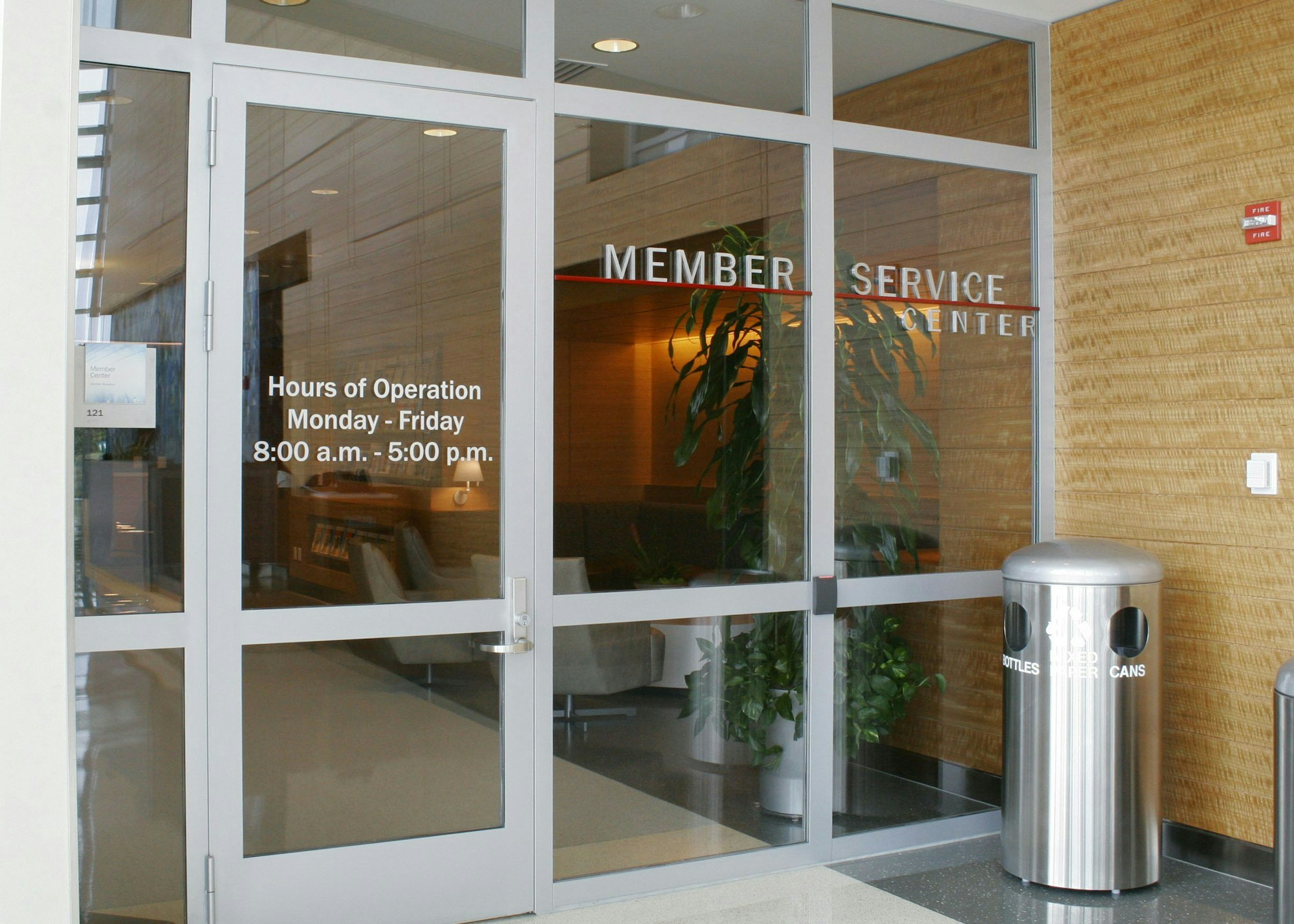Glass doors to a "Member Service Center" with posted hours, an interior view with plants, and recycling bins outside.