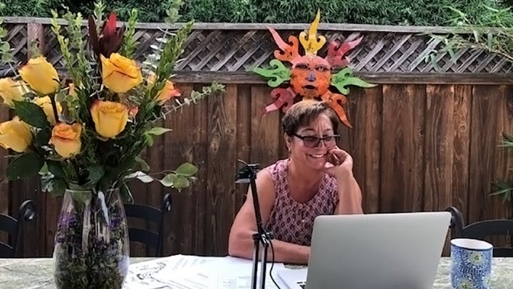 A person smiling at a laptop outside, with a bouquet of yellow roses and a colorful sun decoration on a wooden fence.