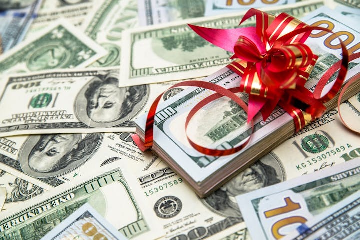 A stack of cash wrapped with a red ribbon sits atop more scattered money. It's a festive presentation of currency.
