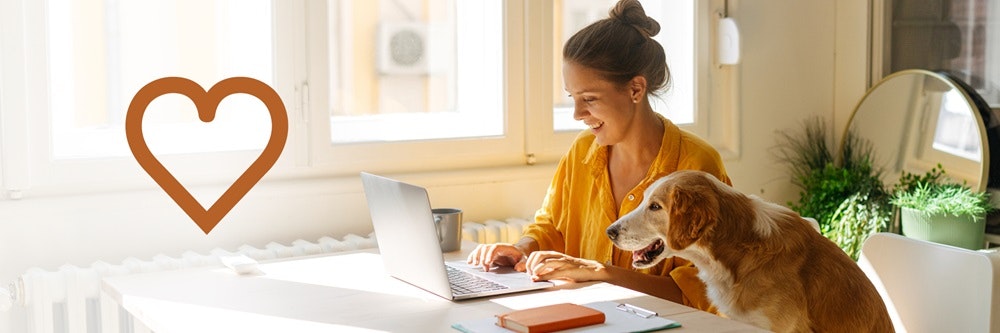 A woman working on a laptop with a dog looking on, and a decorative heart symbol next to them.