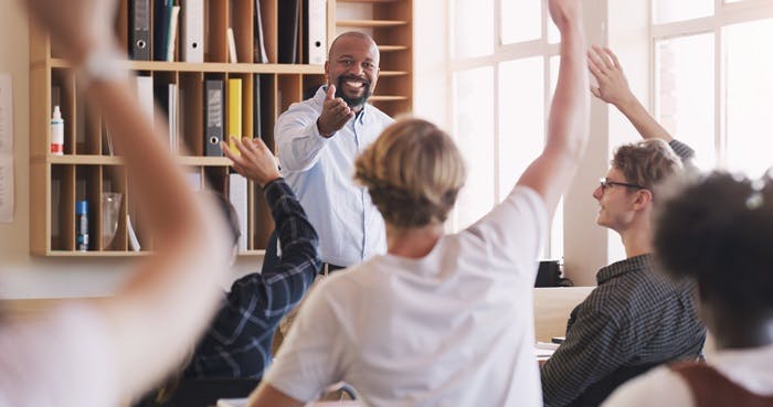 A teacher smiling and pointing at students with raised hands in a classroom.