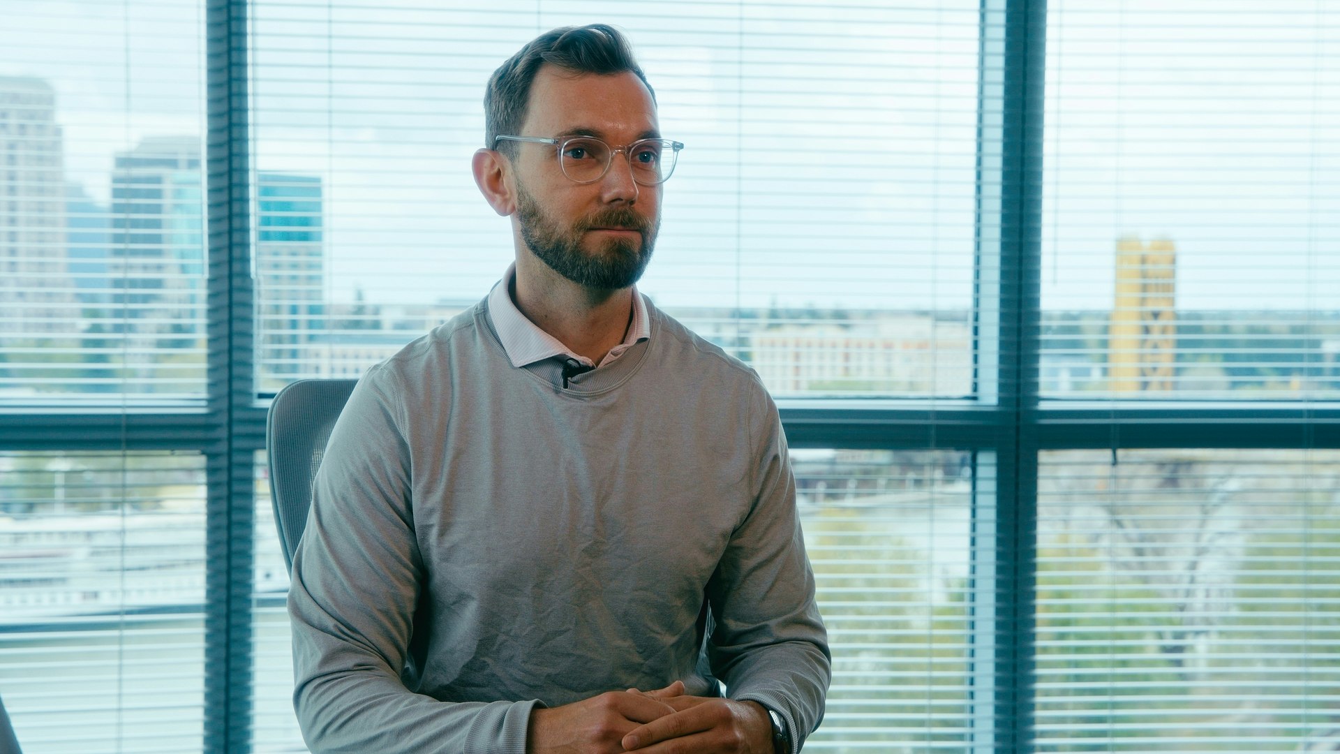 A seated person wearing glasses, in front of a large window with a cityscape in the background.