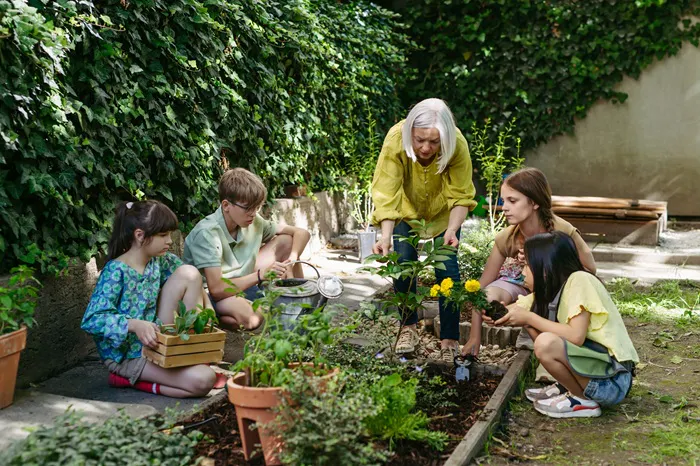 A group of children and an elderly woman are gardening together in a lush green space, tending to plants and flowers.