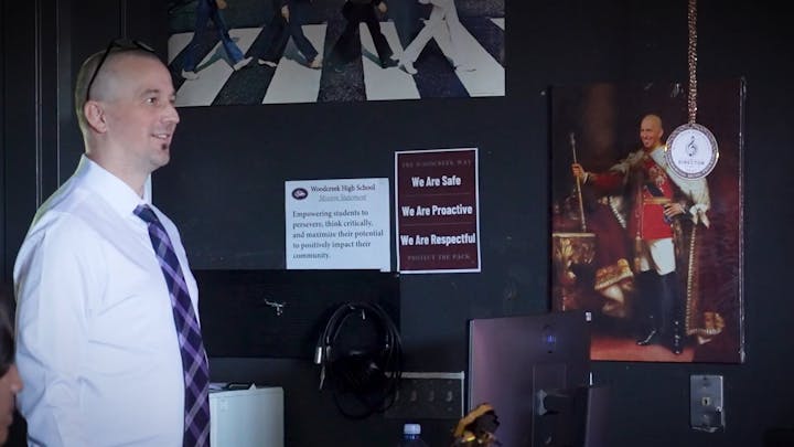 A man in a tie is smiling indoors. Posters and a portrait are on the wall behind him, featuring motivational text and art.