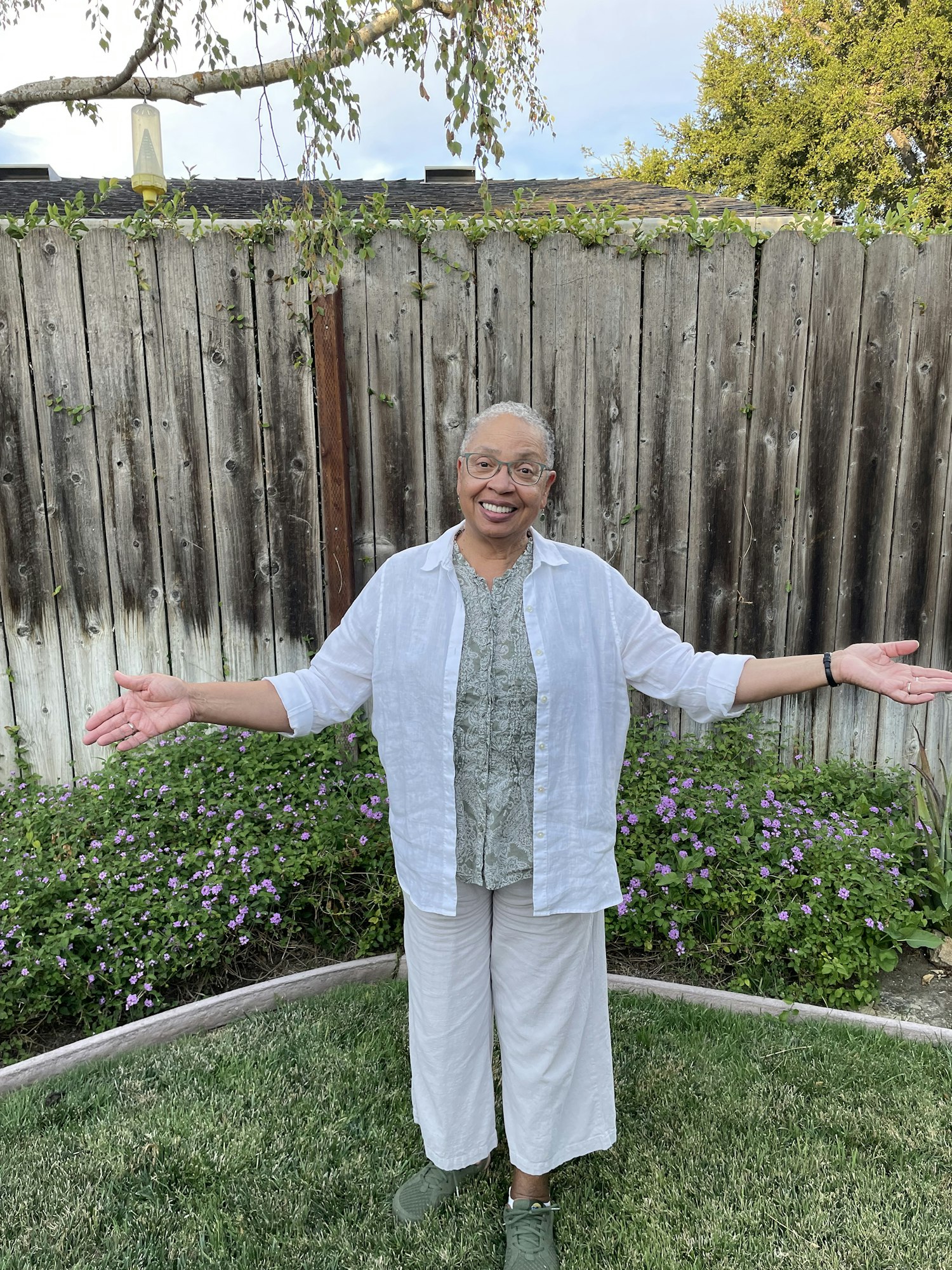 Deborah wears a white shirt while holding her arms out and smiling in her backyard.