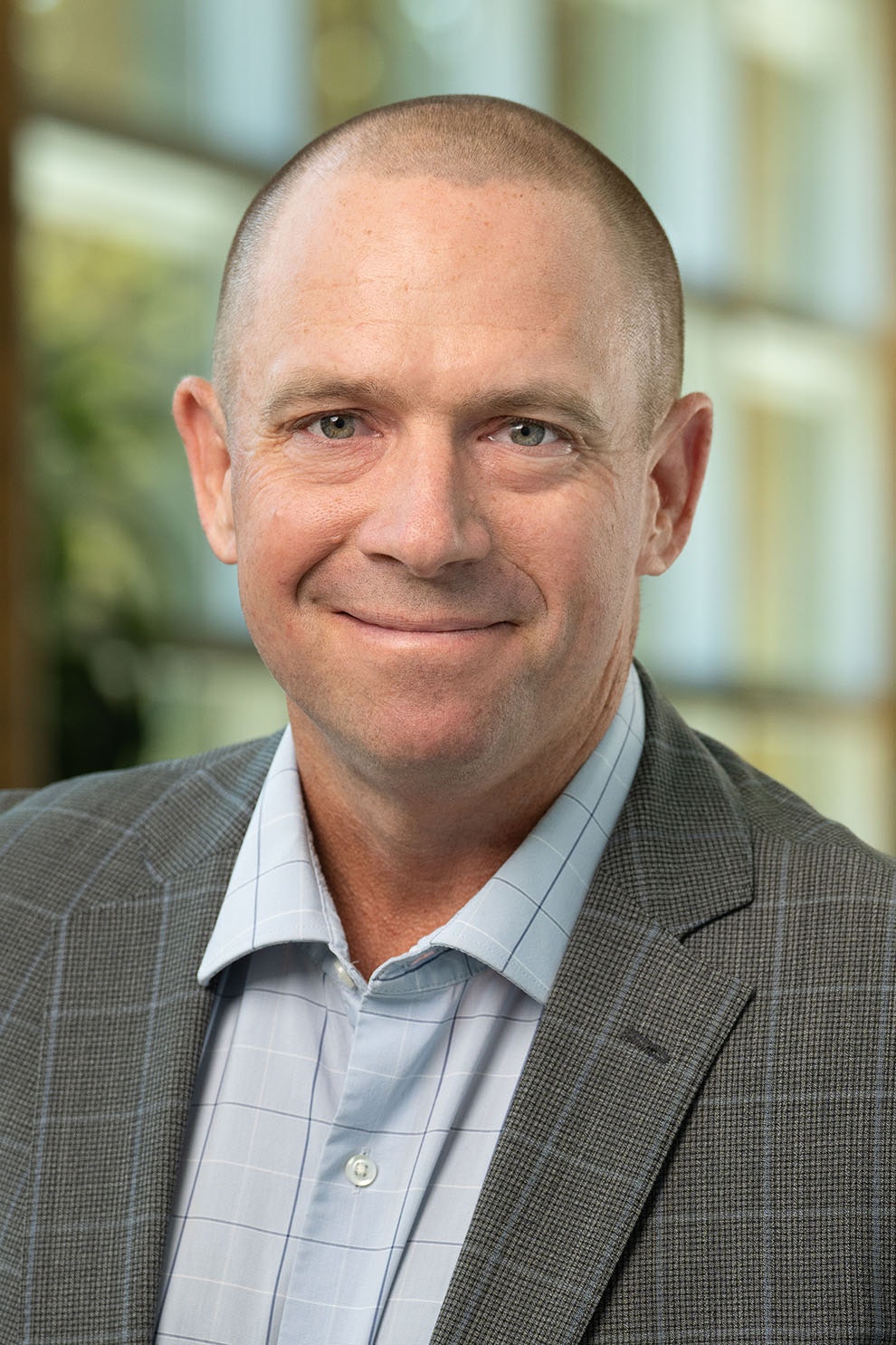 The image shows a smiling man with a shaved head, wearing a plaid blazer and a light blue shirt, in an indoor setting.