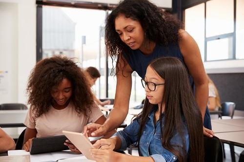 Young teacher with curly brown hair works with students using tablets in a classroom.