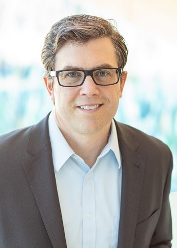 A smiling person with glasses, wearing a suit and a blue shirt, in front of a bright background.