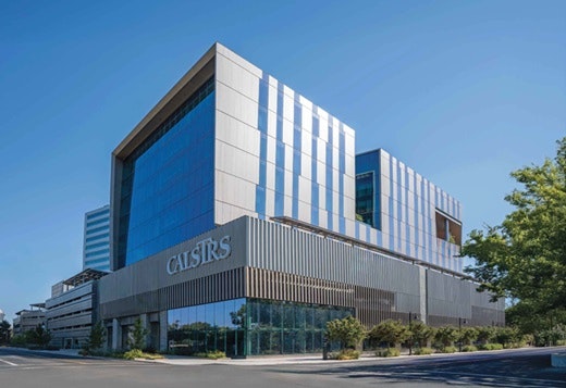 Modern multi-story building with "CalSTRS" sign, glass facade, clear sky, and greenery in front.