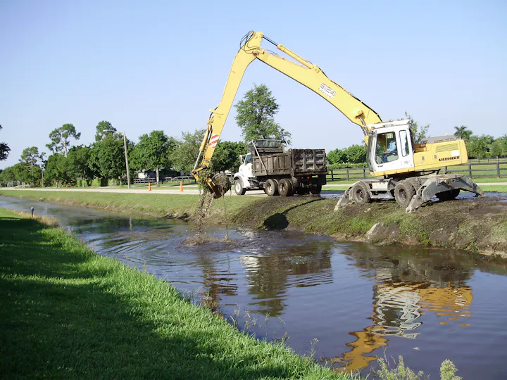 A yellow excavator is removing debris from a canal, with a truck nearby and a green landscape in the background.