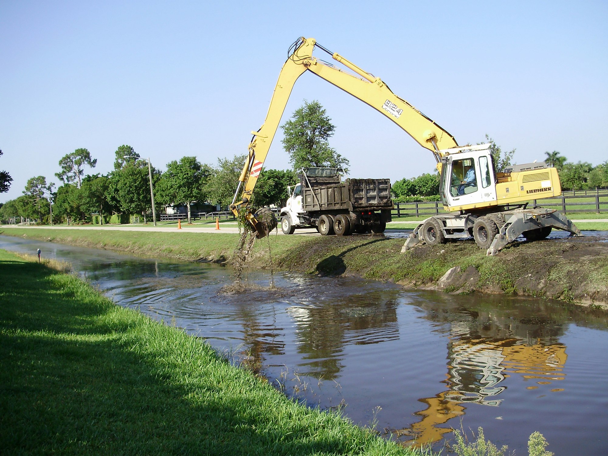 A yellow excavator is removing debris from a canal while a truck waits nearby, surrounded by trees and a clear sky.