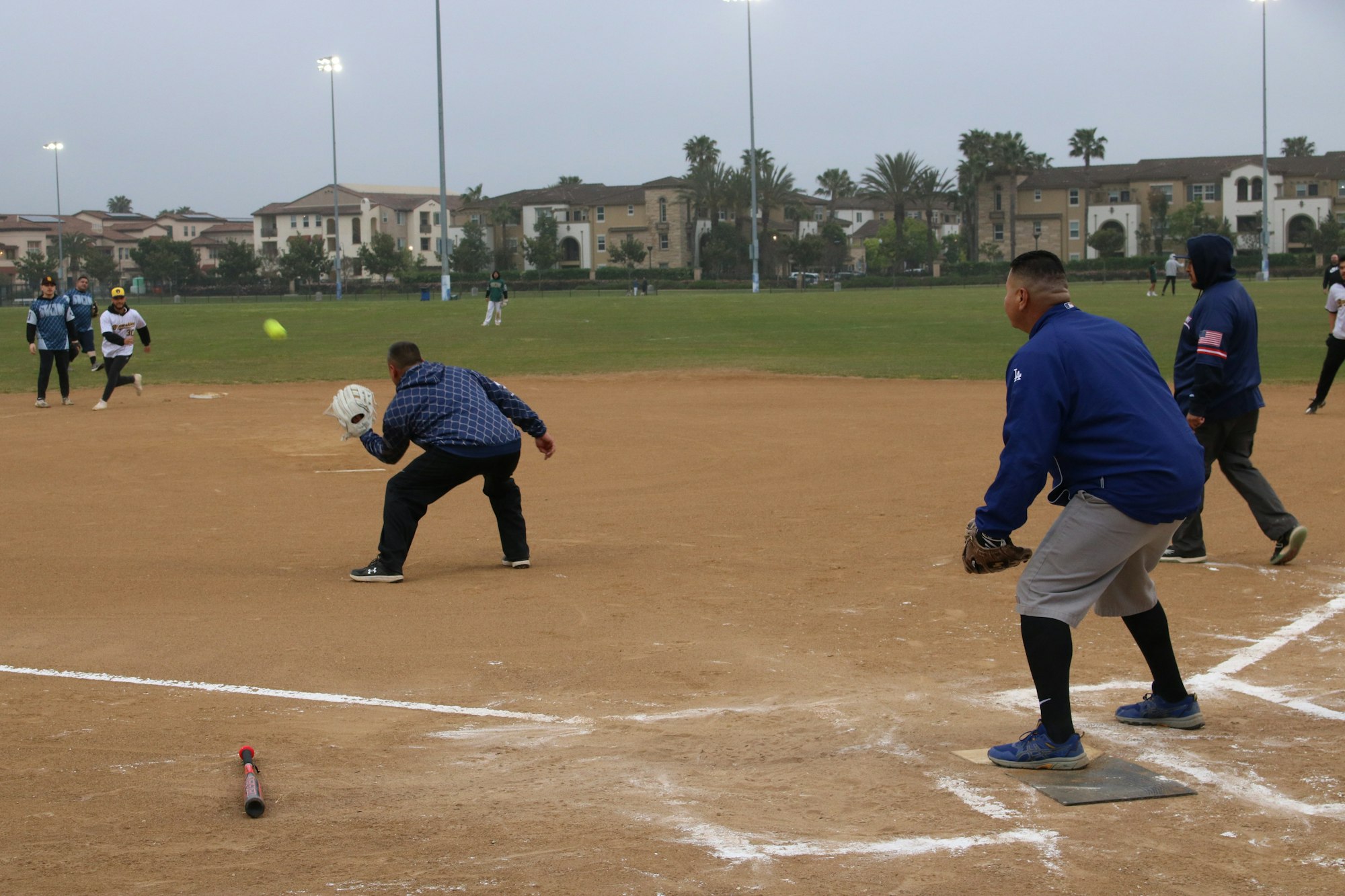 A softball game in progress with players on a dirt field, a bat on the ground, and housing in the background.