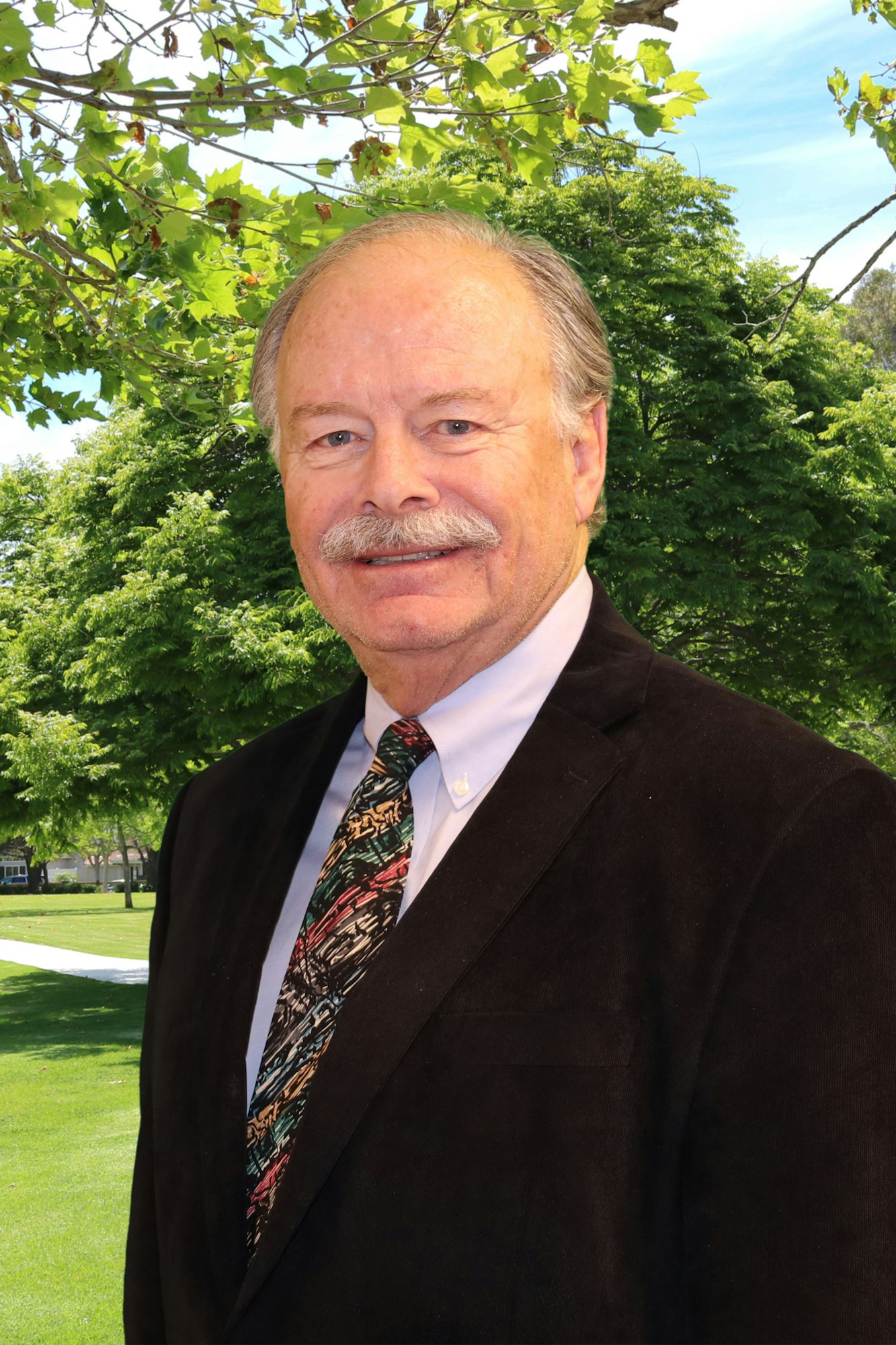 A man in a suit and tie smiles against a backdrop of green trees under a clear blue sky.
