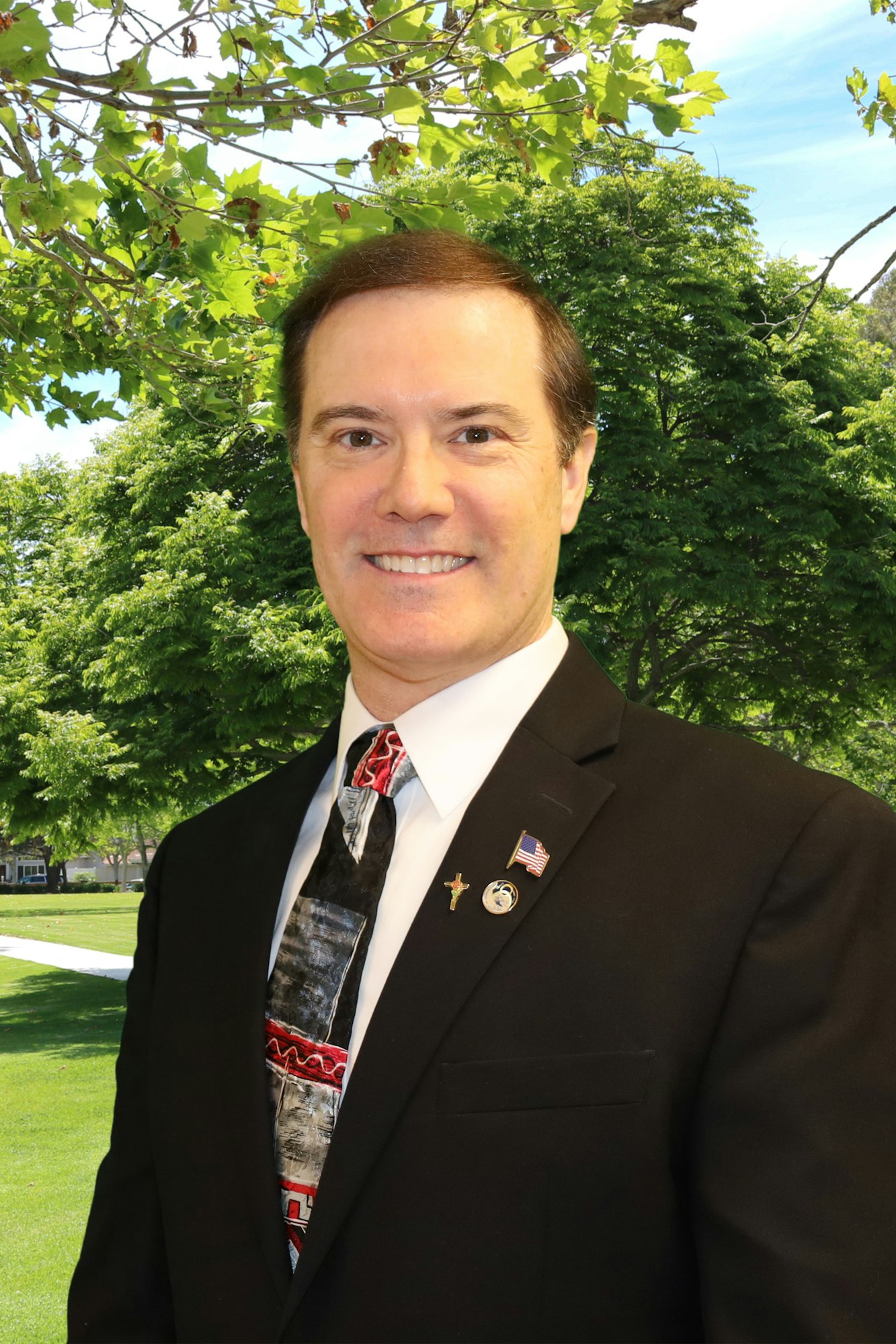 A man in a suit with pins, standing outdoors with trees in the background.