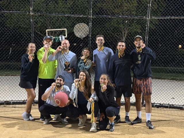 A group of people celebrating with medals and a trophy after a kickball game, posing outdoors at night.