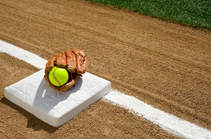 A baseball glove and ball on a white base with a dirt infield and grass outfield.