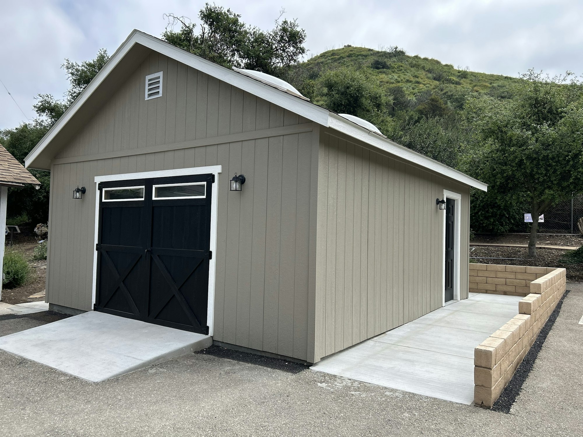 A tan shed with black doors, concrete ramp, and a backdrop of greenery on a hillside.