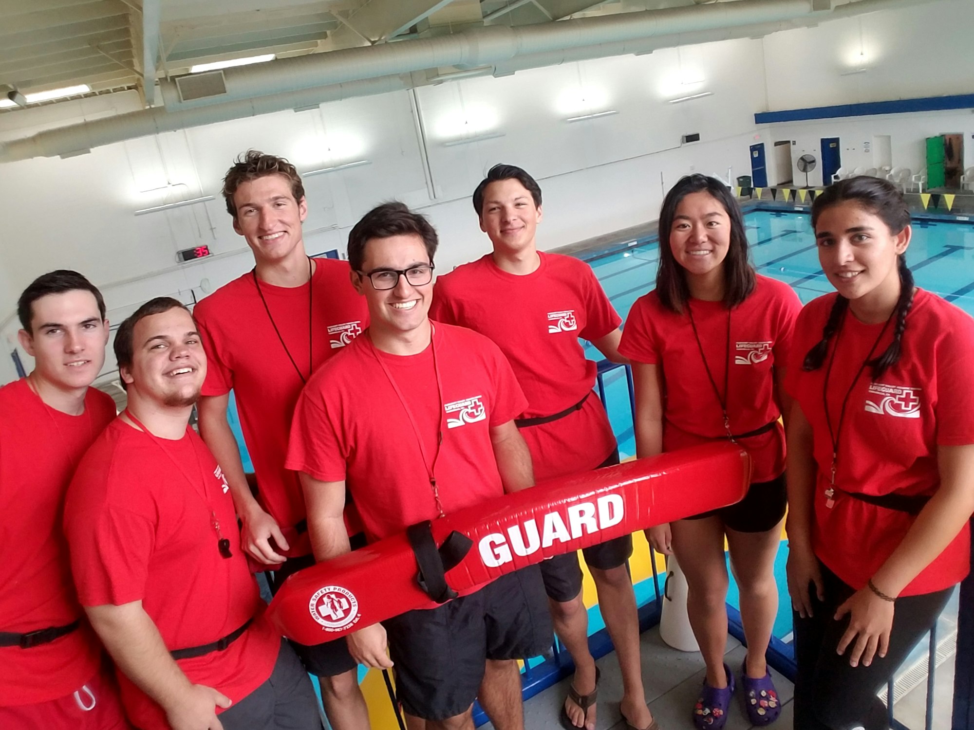 Group of lifeguards in red t-shirts next to a pool, holding a rescue tube labeled "GUARD."