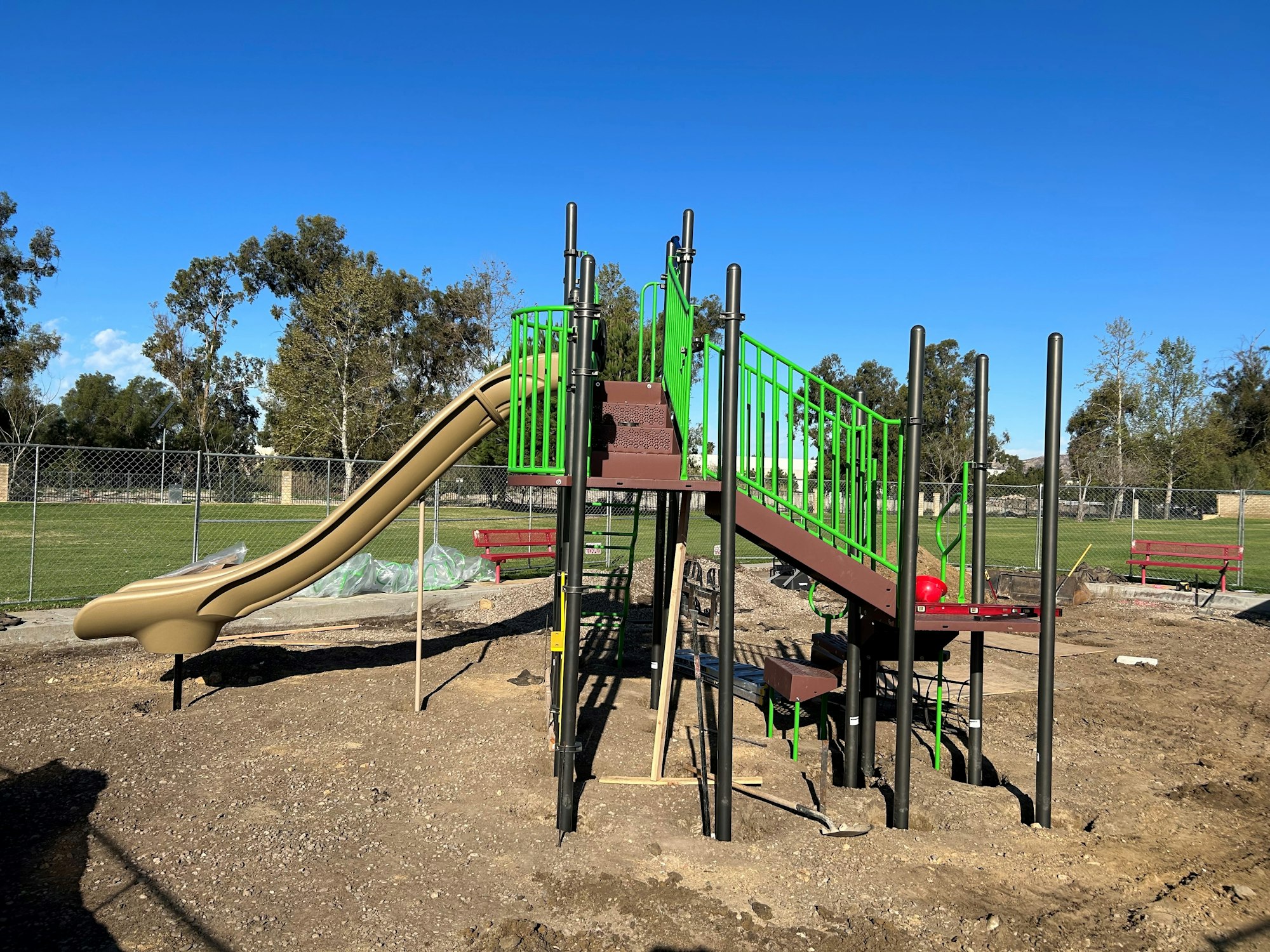 A playground under construction with a slide, climbing steps, and frames. There are red benches and some green grass in the background.
