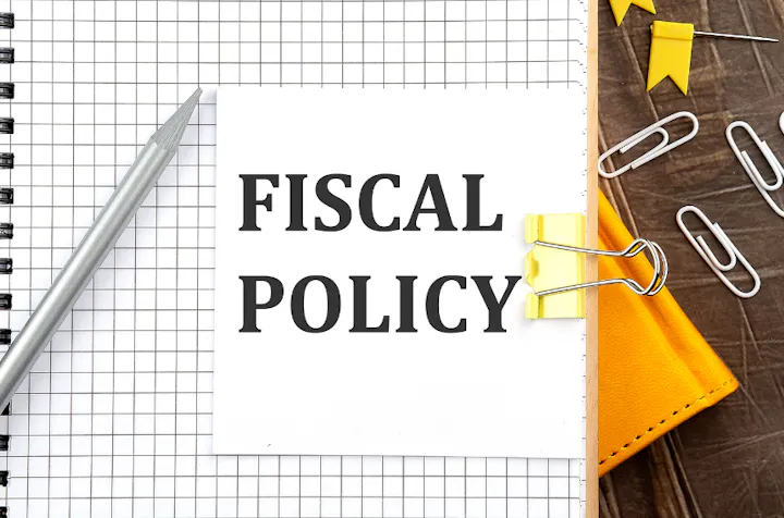 A notepad with "FISCAL POLICY" text, a pencil, paper clips, and a yellow leather case on a wooden surface.