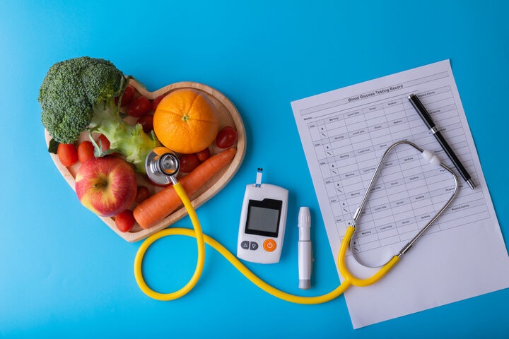 Heart-shaped tray with fruits and veggies, stethoscope, glucometer, pen, and blood glucose record on a blue background.
