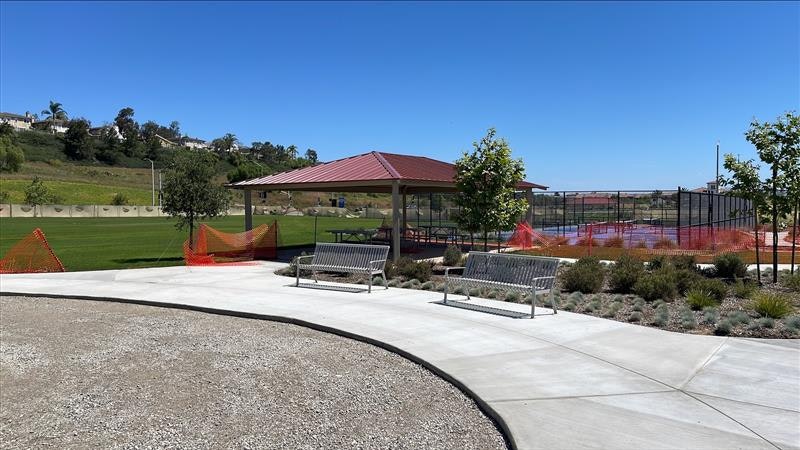 A park scene featuring a pavilion, benches, grass, and construction barriers with trees in the background under a clear blue sky.