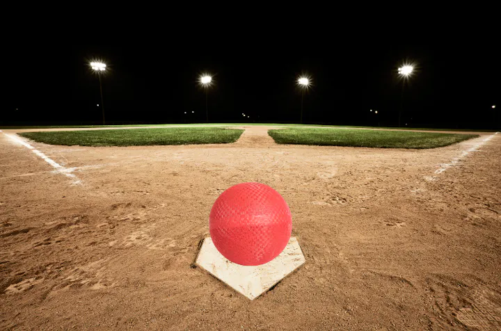 A red kickball on a base of a lit, empty baseball diamond at night.