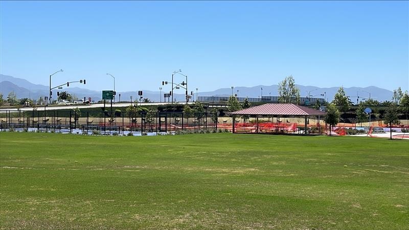 A grassy area with a pavilion, fenced sports courts, a road, and distant mountains under a clear blue sky.