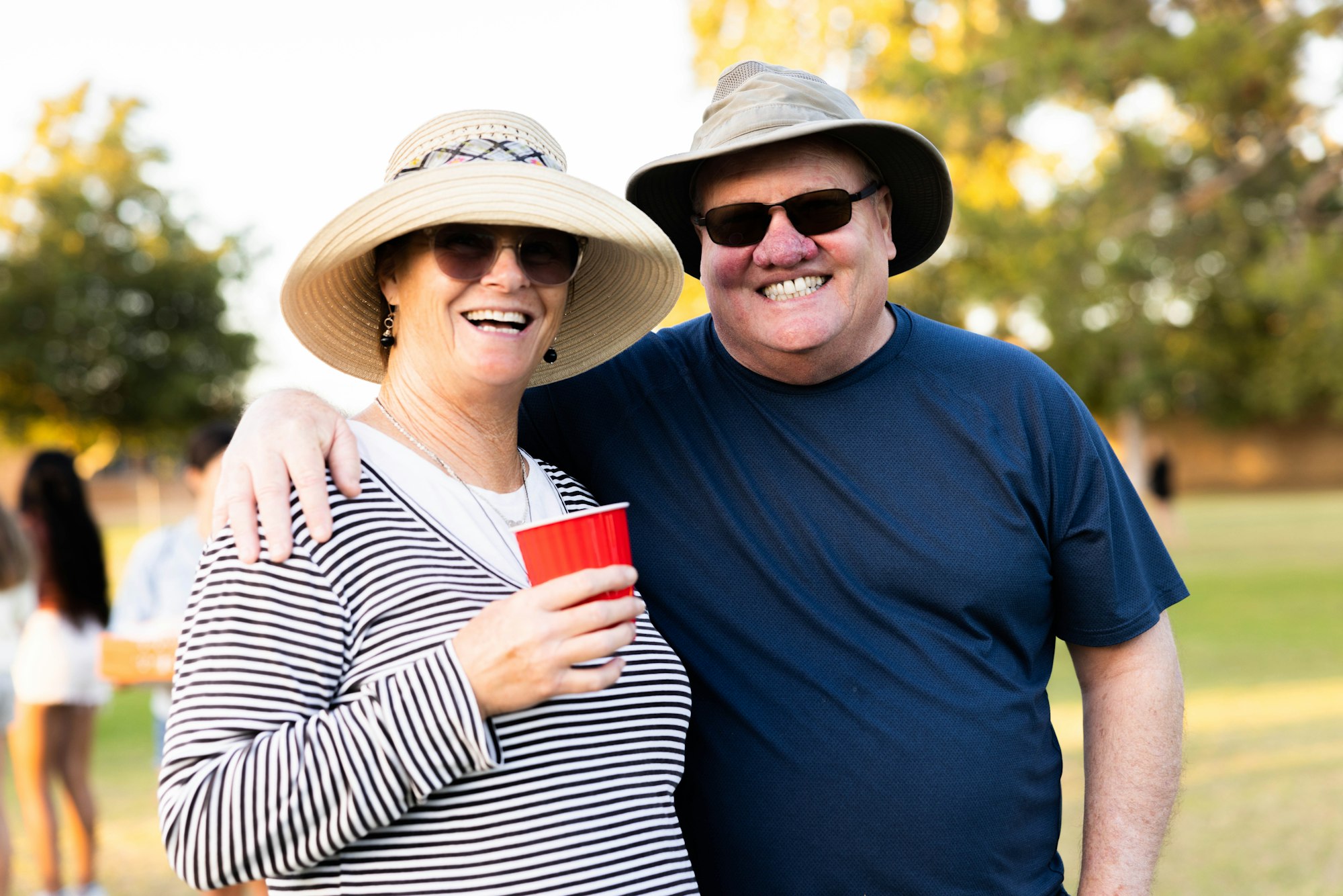 May contain: clothing, hat, sun hat, face, head, person, photography, portrait, t-shirt, accessories, sunglasses, happy, smile, glasses, cap, cup, disposable cup, grass, plant, jewelry, necklace, and laughing