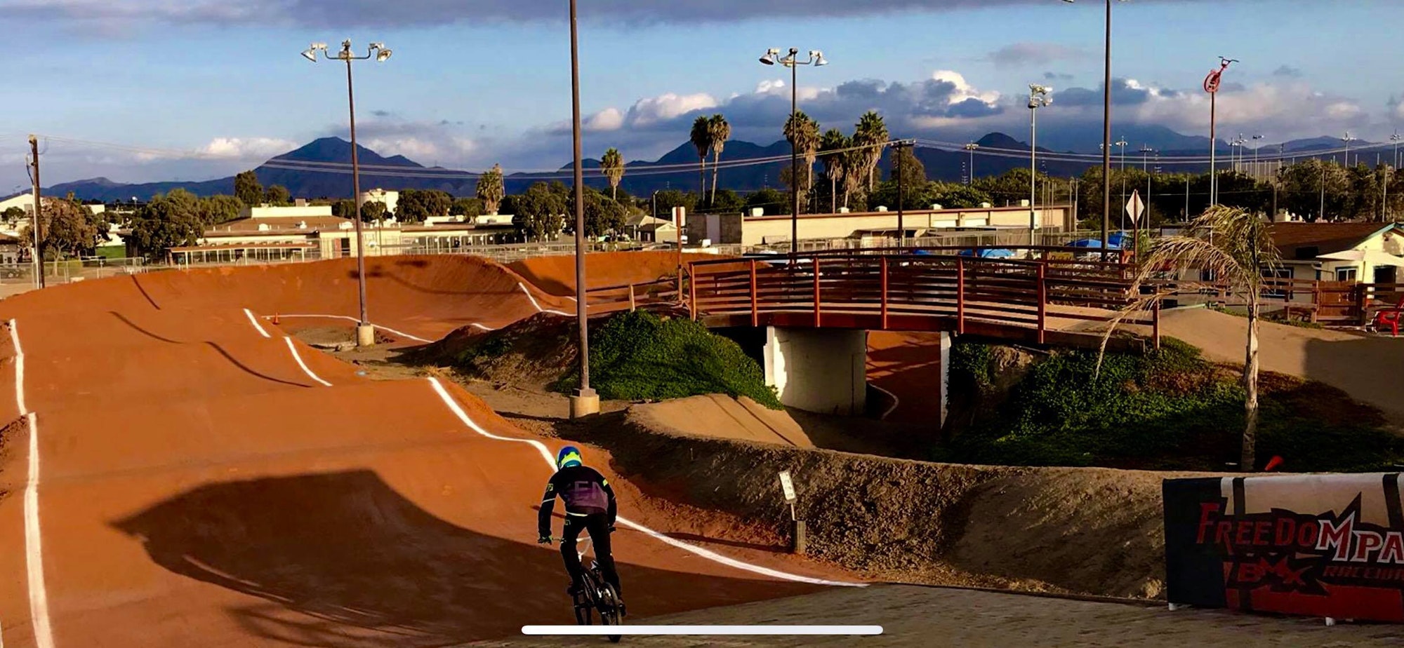 A BMX rider on a dirt track with a bridge in the background and mountains under a partly cloudy sky.
