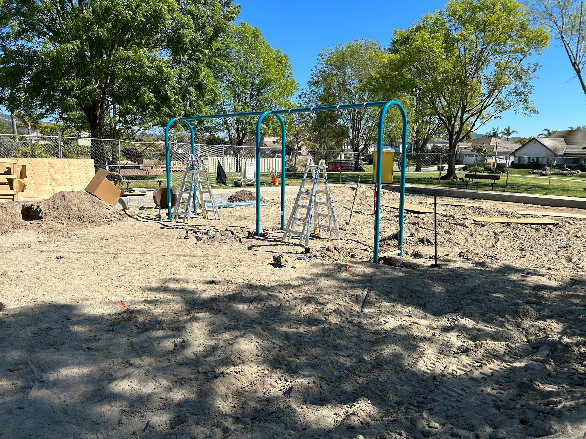 A playground area under construction with ladders and equipment, surrounded by trees and houses.