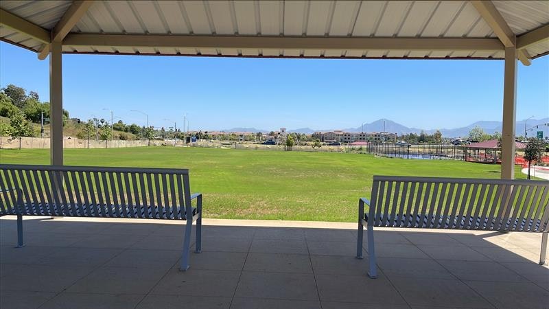 A shaded outdoor space featuring two benches overlooking a green field and distant mountains. Clear blue sky above.