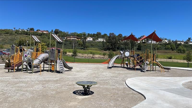 A sunny playground with slides, climbing structures, and shaded areas, set against a green hillside and blue sky.