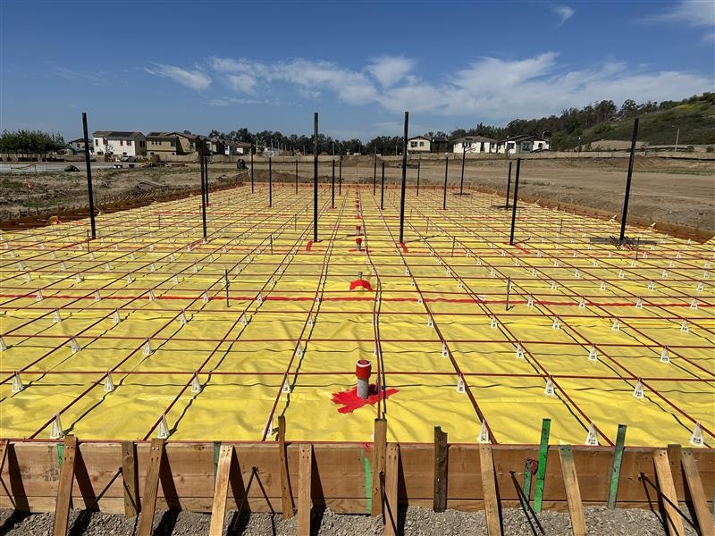 Construction site with yellow sheeting, grid of metal rods, wooden framework, and pipes, against a backdrop of houses and blue sky.