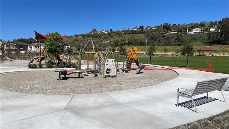 A playground featuring swings, slides, and climbing structures, surrounded by green grass and a clear blue sky.