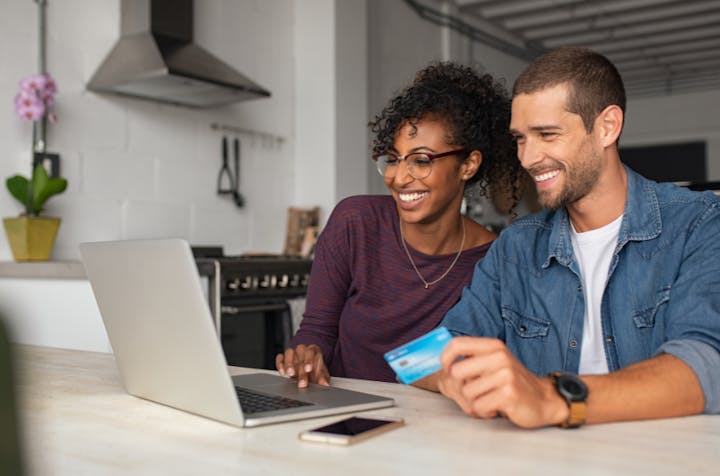 Two people smiling at a laptop, one holding a credit card, likely shopping online.