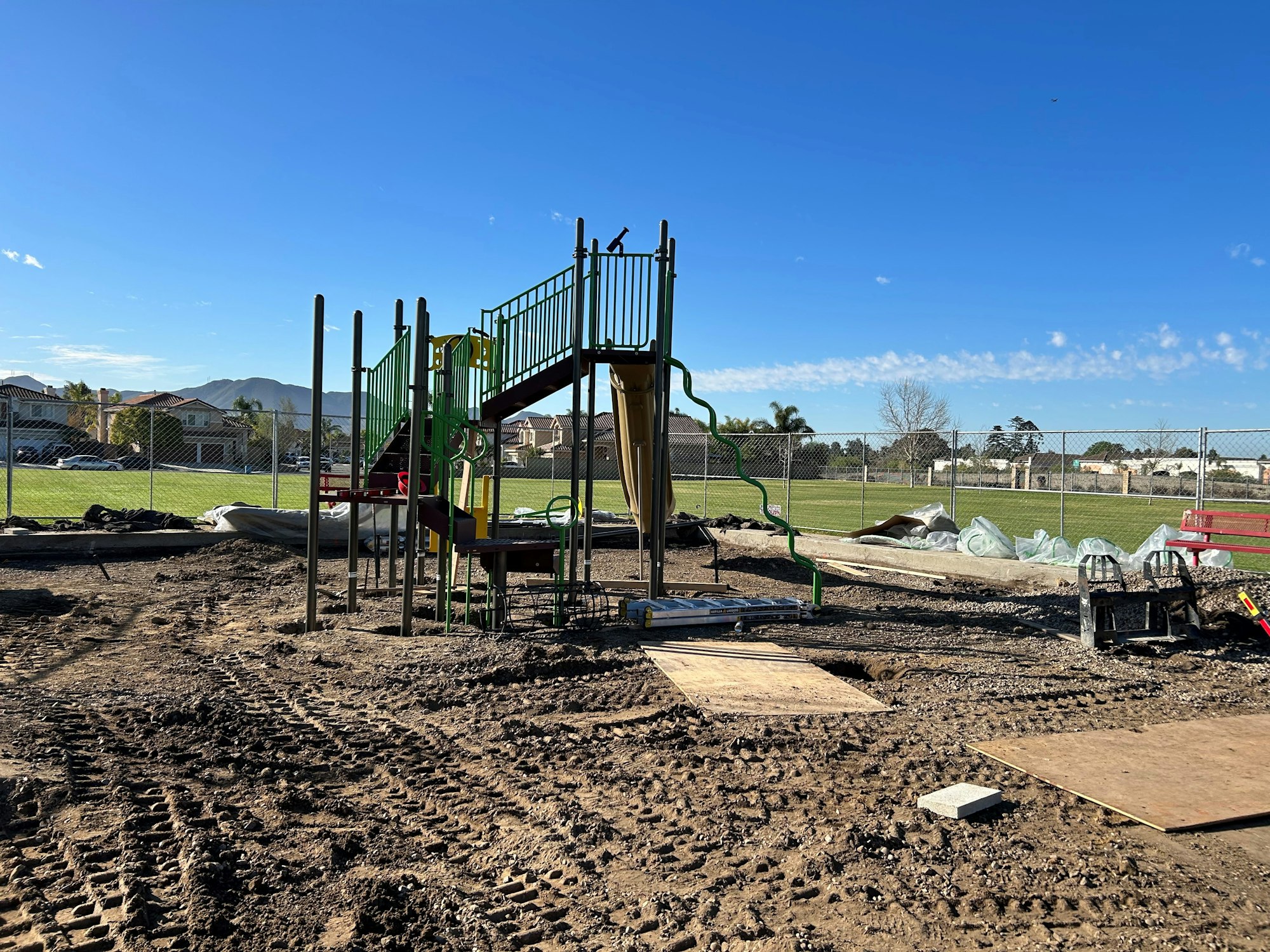 A playground under construction with slides and equipment, surrounded by dirt and a chain-link fence.