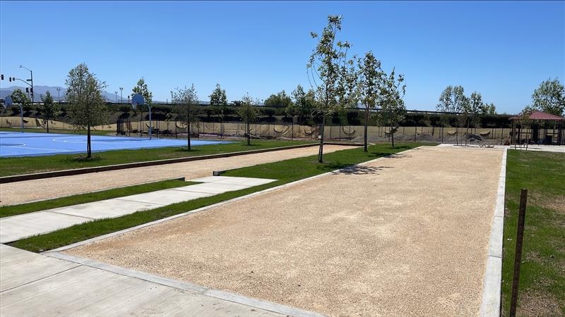 The image shows a park area with a basketball court, walking paths, grass, and newly planted trees.