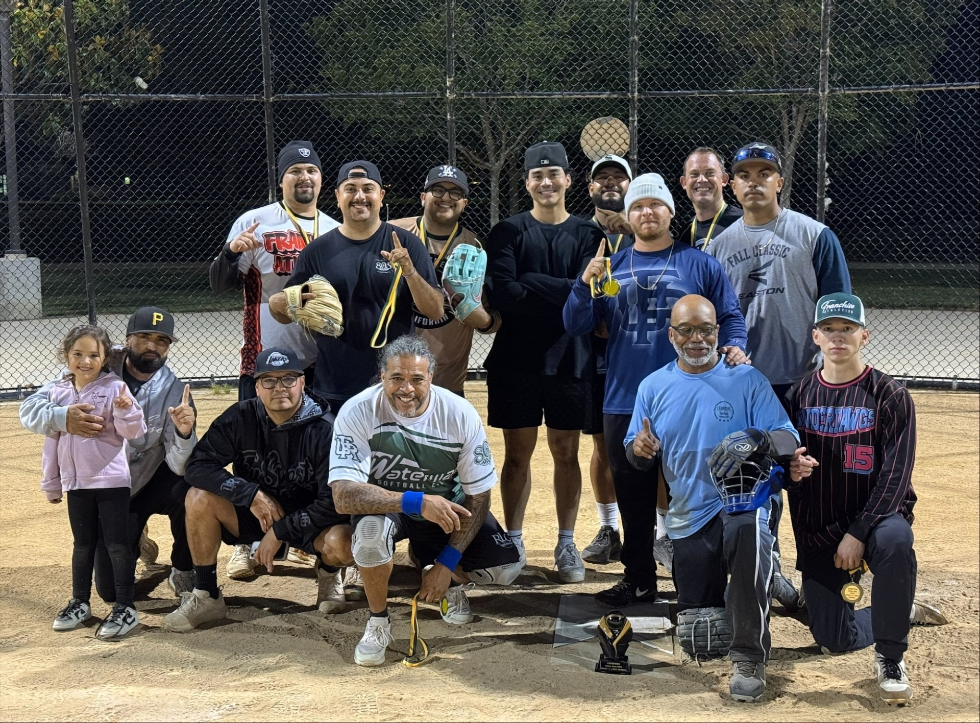 A group of players, including a child, pose after a softball game, celebrating with medals and a trophy at night.