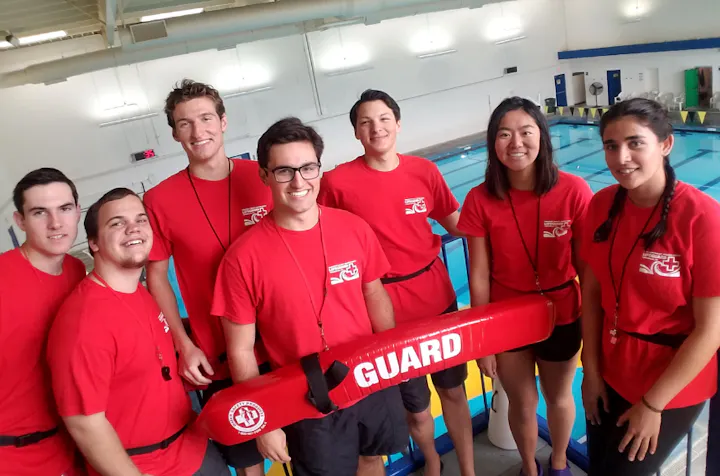 Group of lifeguards in red shirts by a pool, one holding a rescue tube marked "GUARD."
