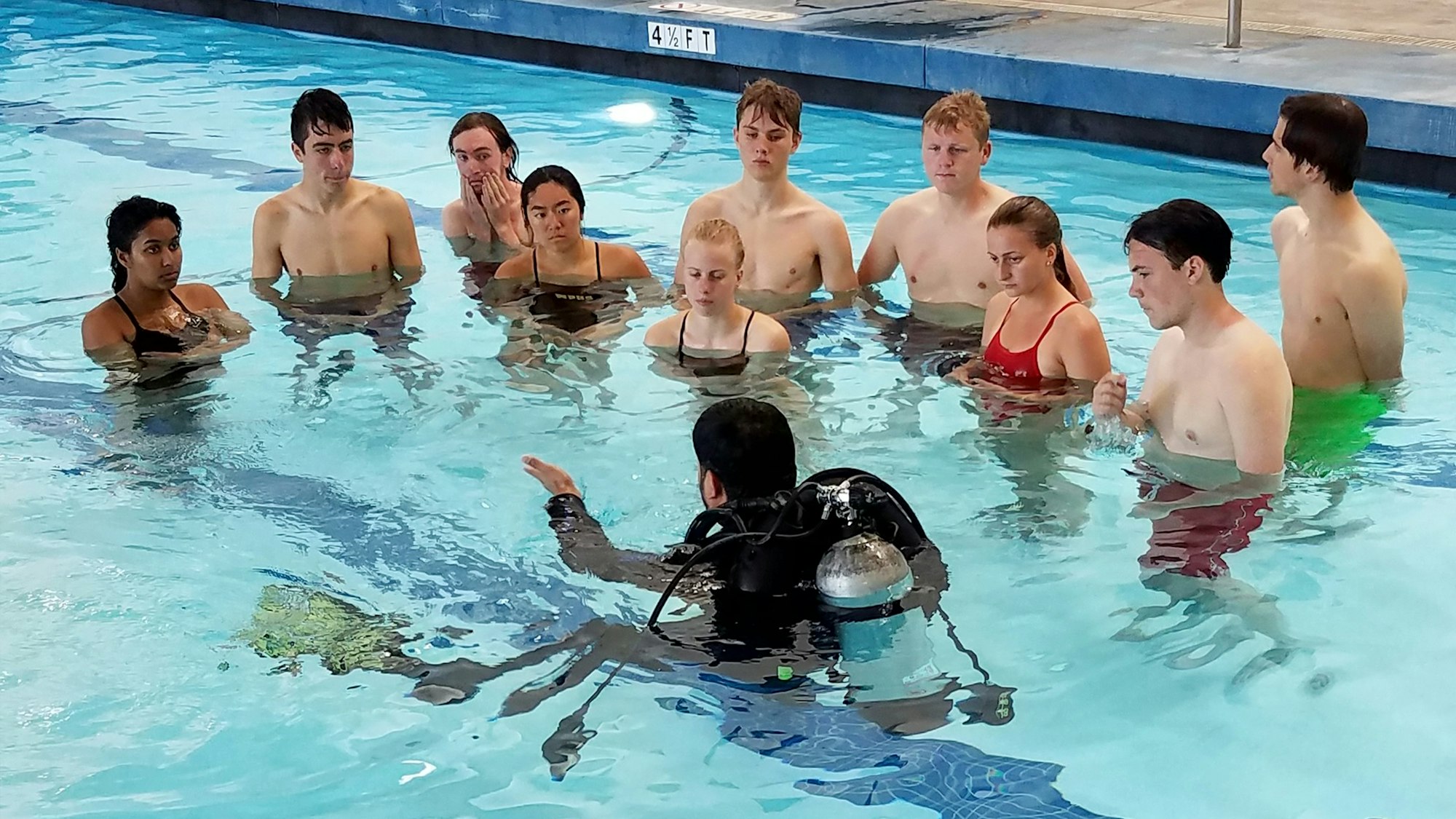 A group of people in a swimming pool receiving instruction from a diver with scuba gear.