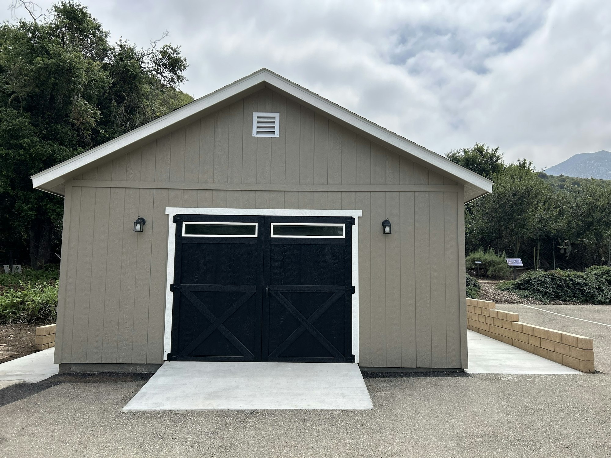 A beige shed with black-trimmed double doors, a vent, outdoor lamps, on a concrete pad with nature in the background.