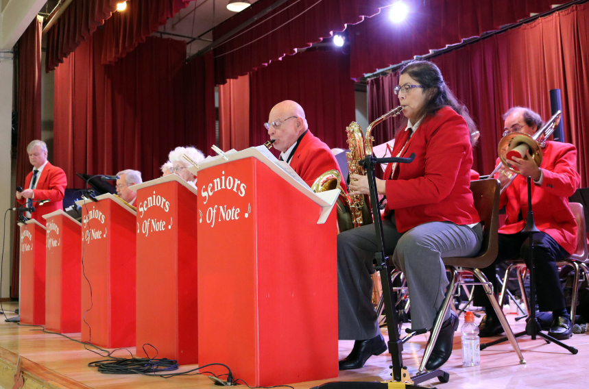 A senior citizens band, "Seniors of Note", performing on stage with red jackets and various instruments.