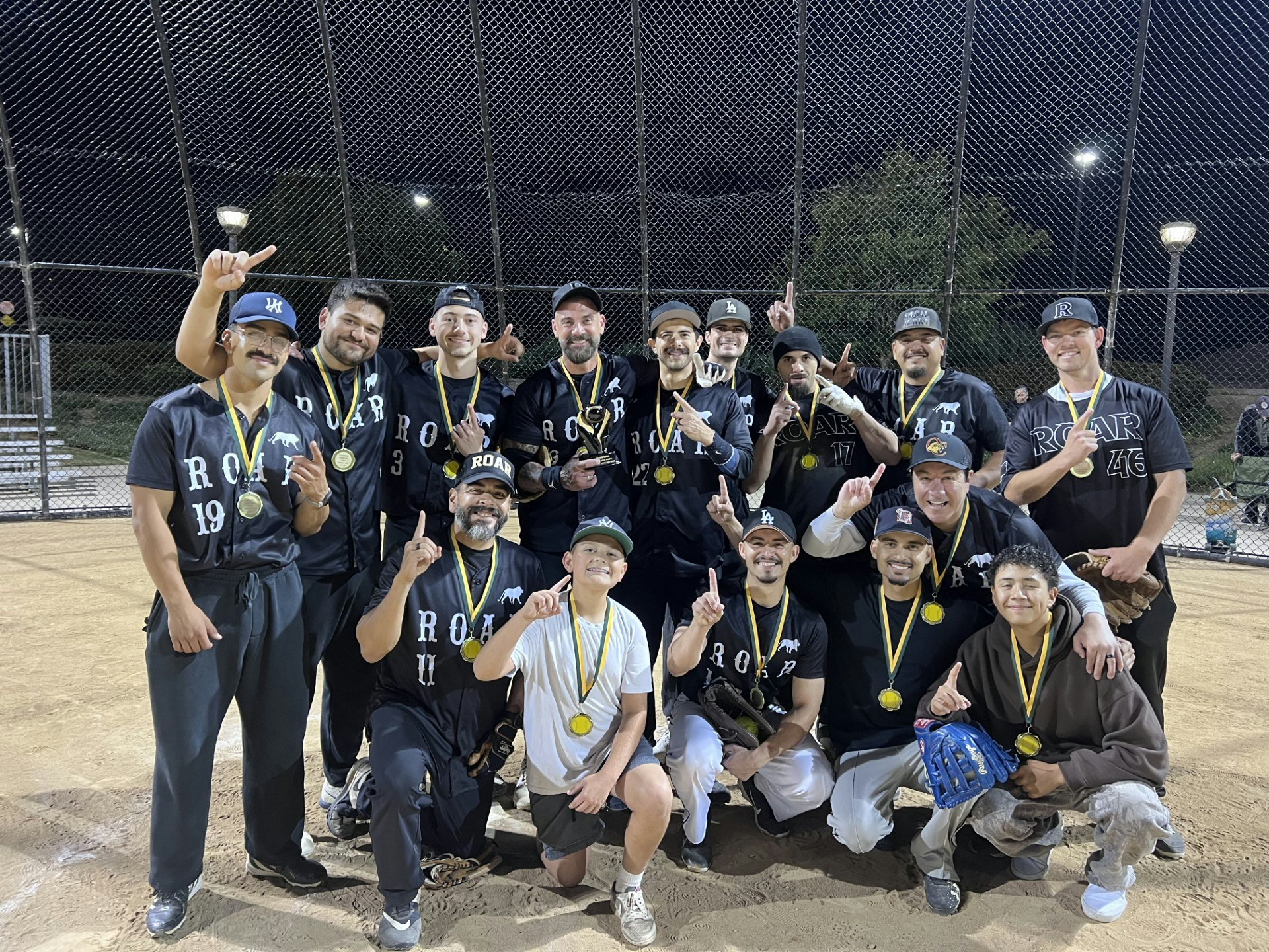 A joyful baseball team poses together on a field, wearing black jerseys and gold medals, celebrating a victory.